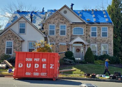 A crew works on the roof of a large stone house covered with blue tarps, with an orange dumpster labeled DUDEZ positioned in front of the house.