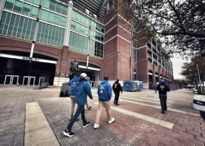 Four people in blue jackets walk toward the entrance of a large stadium with brick walls and green-tinted windows on a cloudy day.