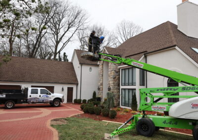 A worker stands on a raised cherry picker near a house, with a company truck parked on the driveway and landscaping visible in the foreground.