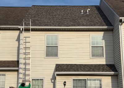 A person in a green shirt stands at the base of a tall ladder leaning against the roof of a beige two-story house on a clear day.