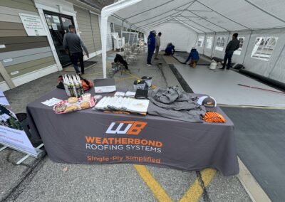 A Weatherbond Roofing Systems table displays brochures, merchandise, and snacks at an outdoor event; people work on roofing materials under a tent in the background.