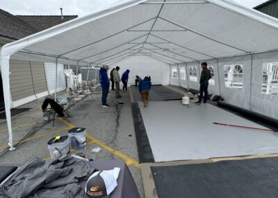 Several people set up flooring under a large white event tent in a parking lot, with tools, chairs, and various supplies scattered around.
