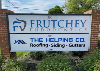 A sign displays two businesses: Frutchey Endodontics at 300 and The Helping Co. Roofing, Siding, Gutters at 306, mounted on a brick frame with plants in front.