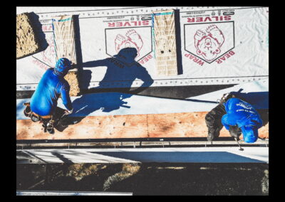 Two construction workers in blue shirts work on a rooftop covered with insulation material labeled Silver Bear, seen from above in bright sunlight.