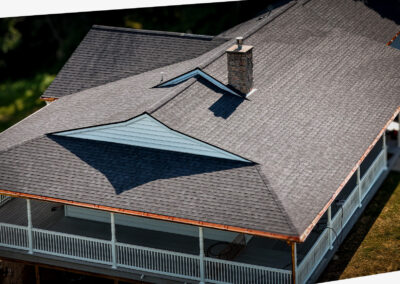 Aerial view of a house with gray, asphalt shingle roofing, a stone chimney, light blue siding, and a wraparound porch.
