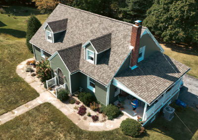 Aerial view of a green house with a gabled roof, dormer windows, front porch, and surrounding garden on a grassy lawn.
