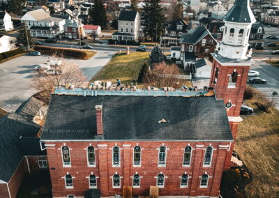 Aerial view of workers repairing the roof of a large brick building with arched windows and a white clock tower in a suburban neighborhood.