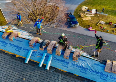 Four workers wearing safety gear install shingles and insulation on a house roof, with equipment and materials scattered on the ground below.