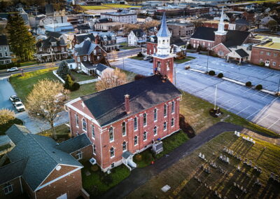 Aerial view of a small town featuring a red-brick church with a tall steeple, a cemetery, surrounding buildings, and mostly empty parking lots.