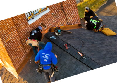 Four workers in safety gear are repairing or installing shingles on a sloped roof next to a brick chimney, using various roofing tools and equipment.