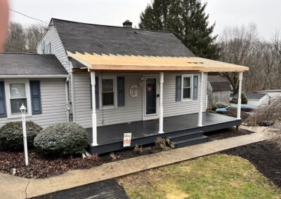 A house with a partially constructed wooden porch roof, white columns, and gray siding on a cloudy day. Construction materials are visible and the yard appears landscaped.