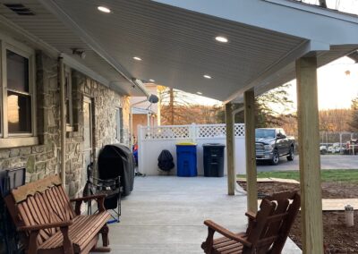 A covered concrete patio with two wooden benches, a grill, trash bins, and a view of a driveway with a parked truck and trees in the background.