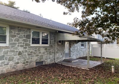 Back view of a stone house with several windows, a small covered patio, fallen leaves on the ground, and a white fence in the background.