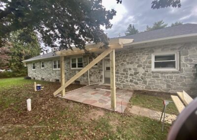 A stone house with a back patio under construction, featuring wooden beams for a future covering and stone tiles on the ground.