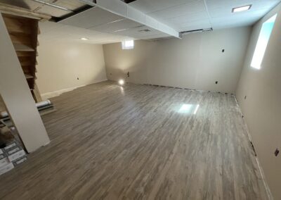 A basement room with newly installed wood-look flooring, beige walls, exposed ceiling panels, and a single light source on the floor.