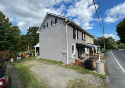 Two-story gray house with a covered front porch, trash bins, and a gravel driveway along a paved road under a partly cloudy sky.