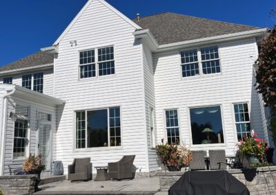 A two-story white house with many windows, outdoor patio furniture, potted plants, and a covered grill on a stone patio under a clear blue sky.