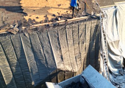 Two workers in blue jackets remove old shingles from a house roof, with tarps covering the walls and a dumpster collecting debris below.