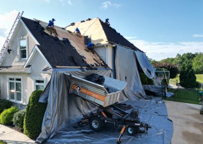 Workers replace the roof of a house, removing old shingles and covering areas with tarps while debris is loaded into a trailer below.