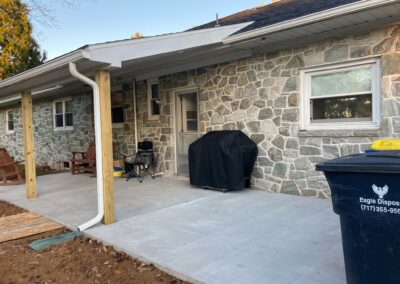 A stone house with a covered patio, two wooden posts, a grill covered with a black cover, a folding chair, and a trash bin labeled Eagle Disposal.