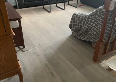 A living room with two black sofas, a patterned armchair, a glass coffee table, and light-colored wood flooring. Some clutter is visible on the coffee table and floor.