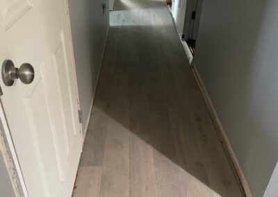 A hallway with newly installed light wood flooring, unpainted baseboards, and doors leading to adjacent rooms. Natural light illuminates part of the floor.