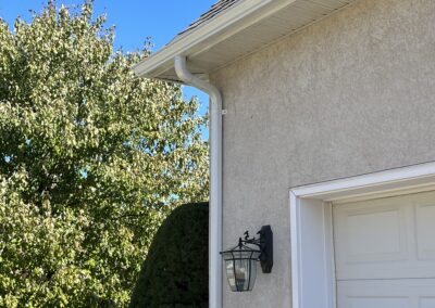 A beige stucco house with white gutters, an outdoor lantern, and a white garage door. Trees and a clear blue sky are visible in the background.