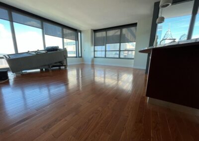 A mostly empty room with large windows, hardwood floors, a gray sofa facing away, and partial view of a dark kitchen counter on the right.