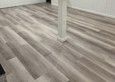 A spacious room with light-colored wood-look vinyl plank flooring, white walls, a central support column, and minimal furniture.