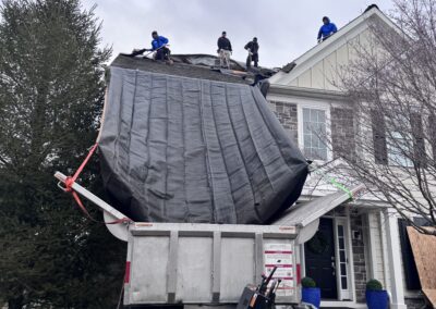 Four workers are on a house roof removing materials, which are being funneled into a dump truck below using a large black tarp chute.