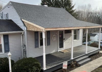 A front view of a house with a covered porch under renovation, indicated by missing fascia and a nearby red dumpster. A sign is placed on the porch steps.