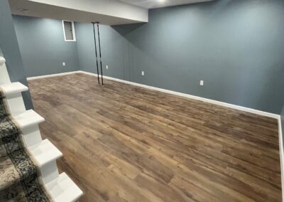 Finished basement room with wood-style flooring, blue-gray walls, recessed lighting, white trim, and stairs with patterned carpet visible in the corner.