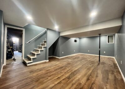 Modern finished basement with wood-look flooring, gray walls, recessed lighting, and a stairway leading up; a support pole is positioned near the middle of the room.