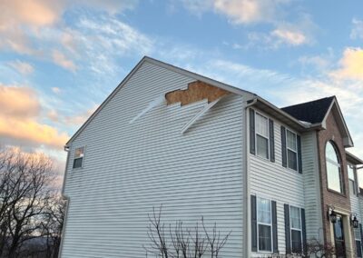 A two-story house with a section of siding missing near the roofline, exposing the underlying sheathing, under a partly cloudy sky.