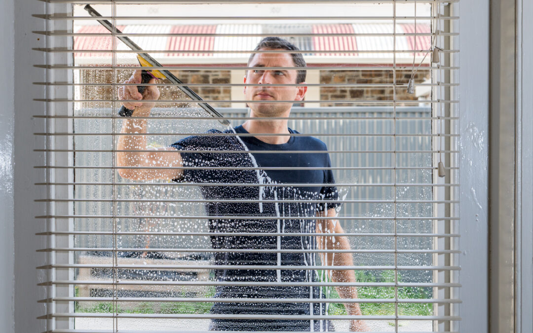 A man uses a squeegee to clean the exterior of a window, visible through partially open blinds with soap and water on the glass.